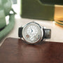 Silver-toned round watch with white dial, circular time display, black leather strap, displayed on brown leather surface.