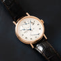 Gold-tone watch with white dial, Roman numerals, multiple subdials, and black leather strap on dark background.