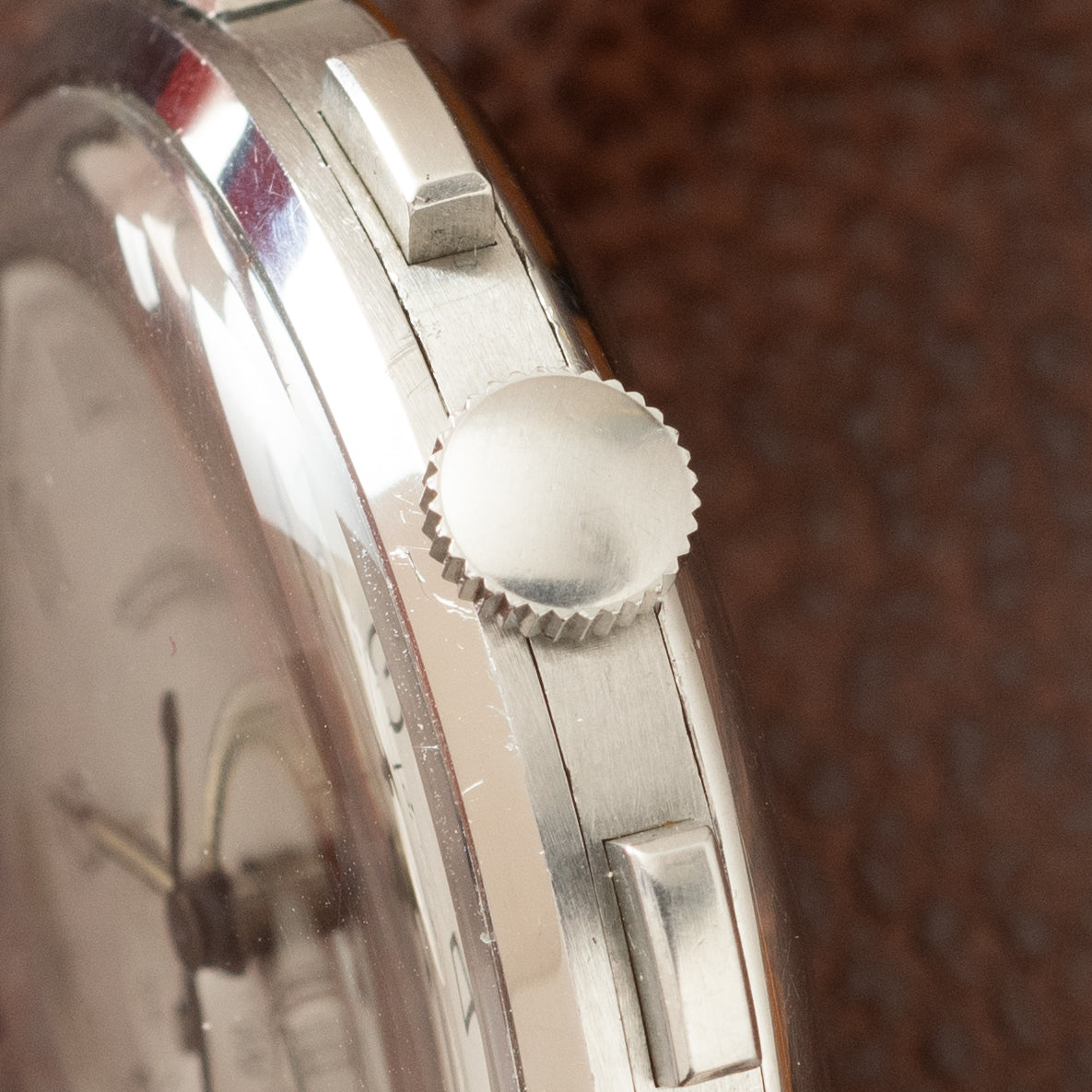 Stainless steel watch case side view showing textured crown, lugs, and brushed metal band against brown background.
