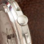Stainless steel watch case side view showing textured crown, lugs, and brushed metal band against brown background.