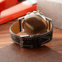 Stainless steel chronograph watch with dark brown leather strap and white stitching, displayed on wooden surface.