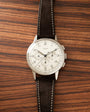 Silver-case chronograph watch with cream dial, black leather strap with white stitching, displayed on wood surface.