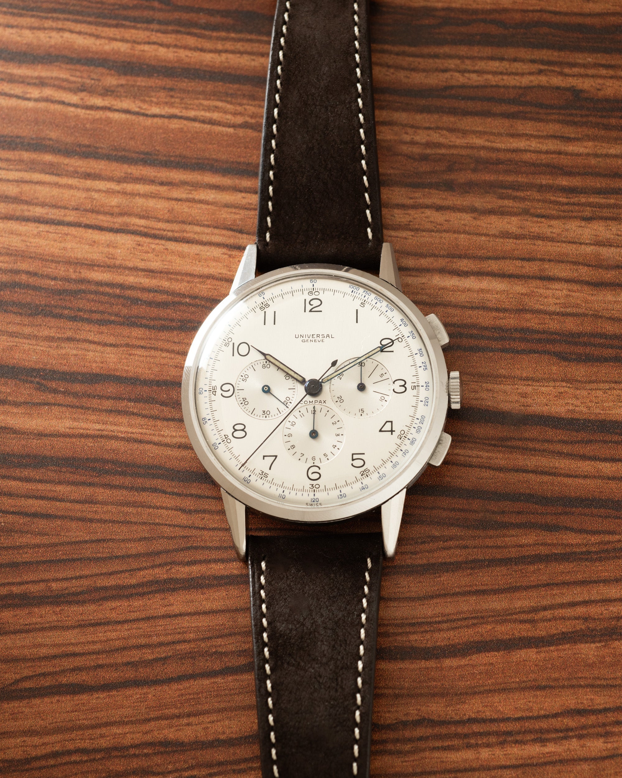 Silver-case chronograph watch with cream dial, black leather strap with white stitching, displayed on wood surface.
