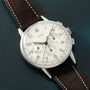 Silver-cased chronograph watch with cream dial, Arabic numerals, two subdials, and dark brown leather strap with white stitch