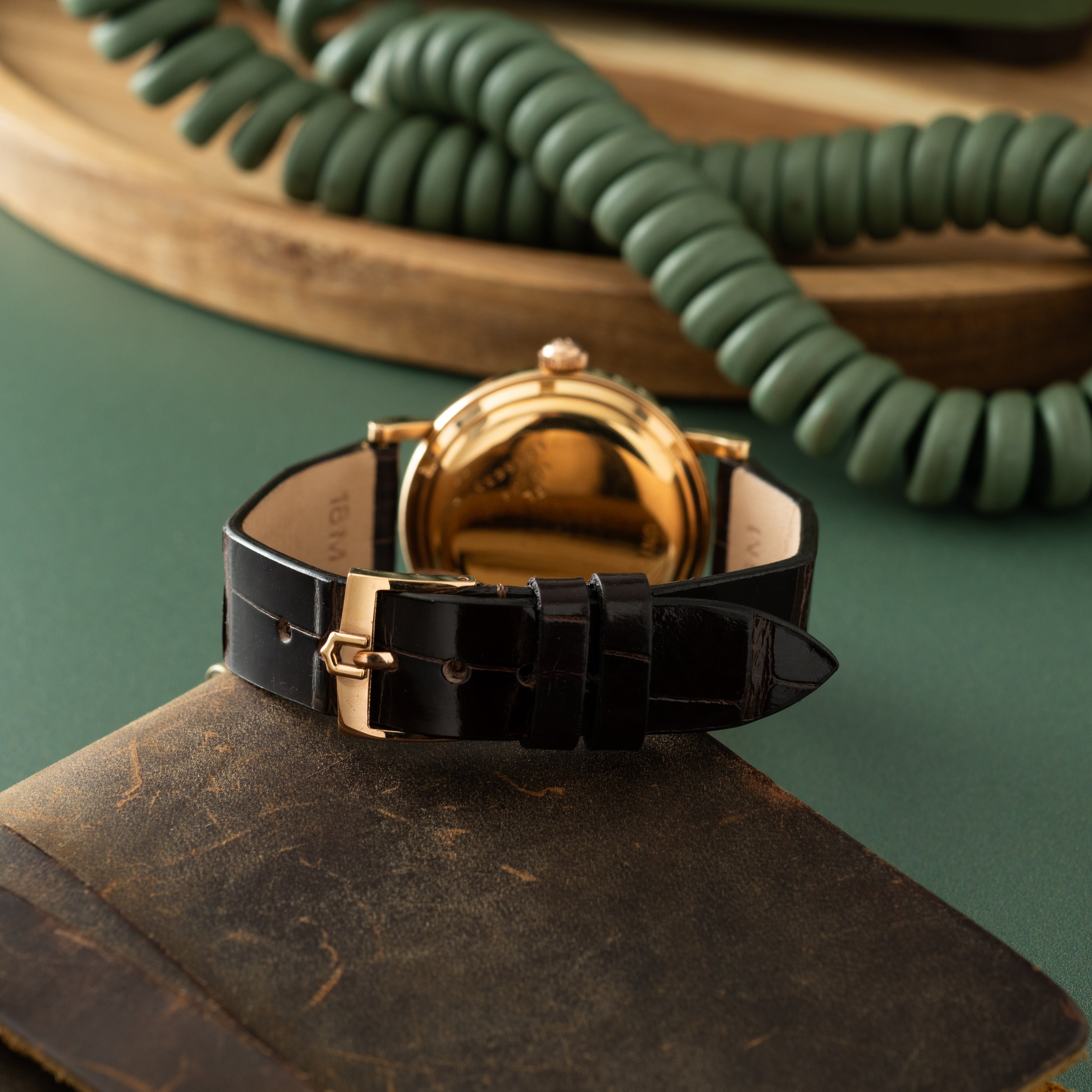 Gold-tone round watch with black leather strap, displayed on vintage book with green coiled phone cord in background.