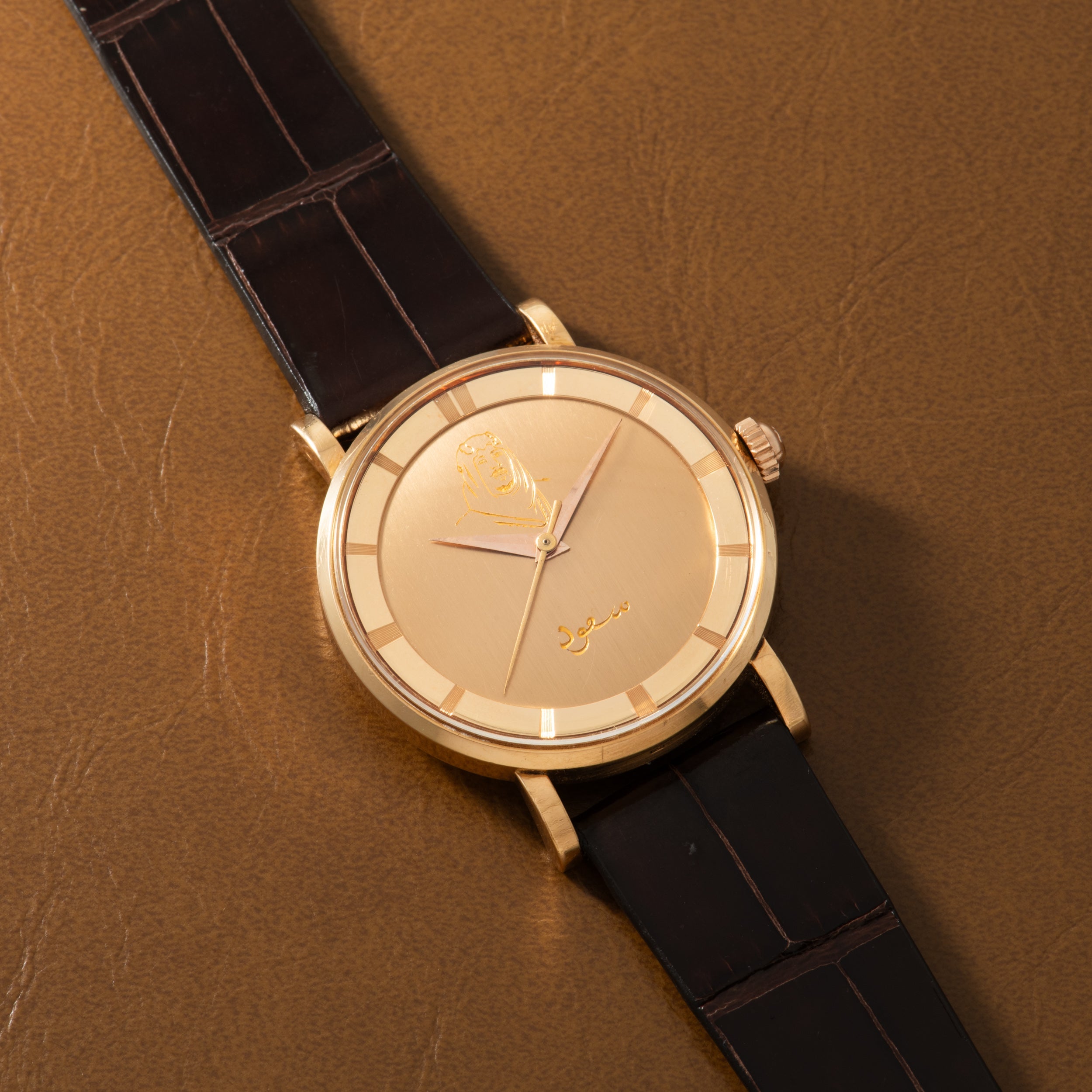 Gold-toned round watch with cream dial, applied indices, and black crocodile leather strap on tan background.