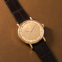 Gold-toned round watch with cream dial, applied indices, and black crocodile leather strap on tan background.