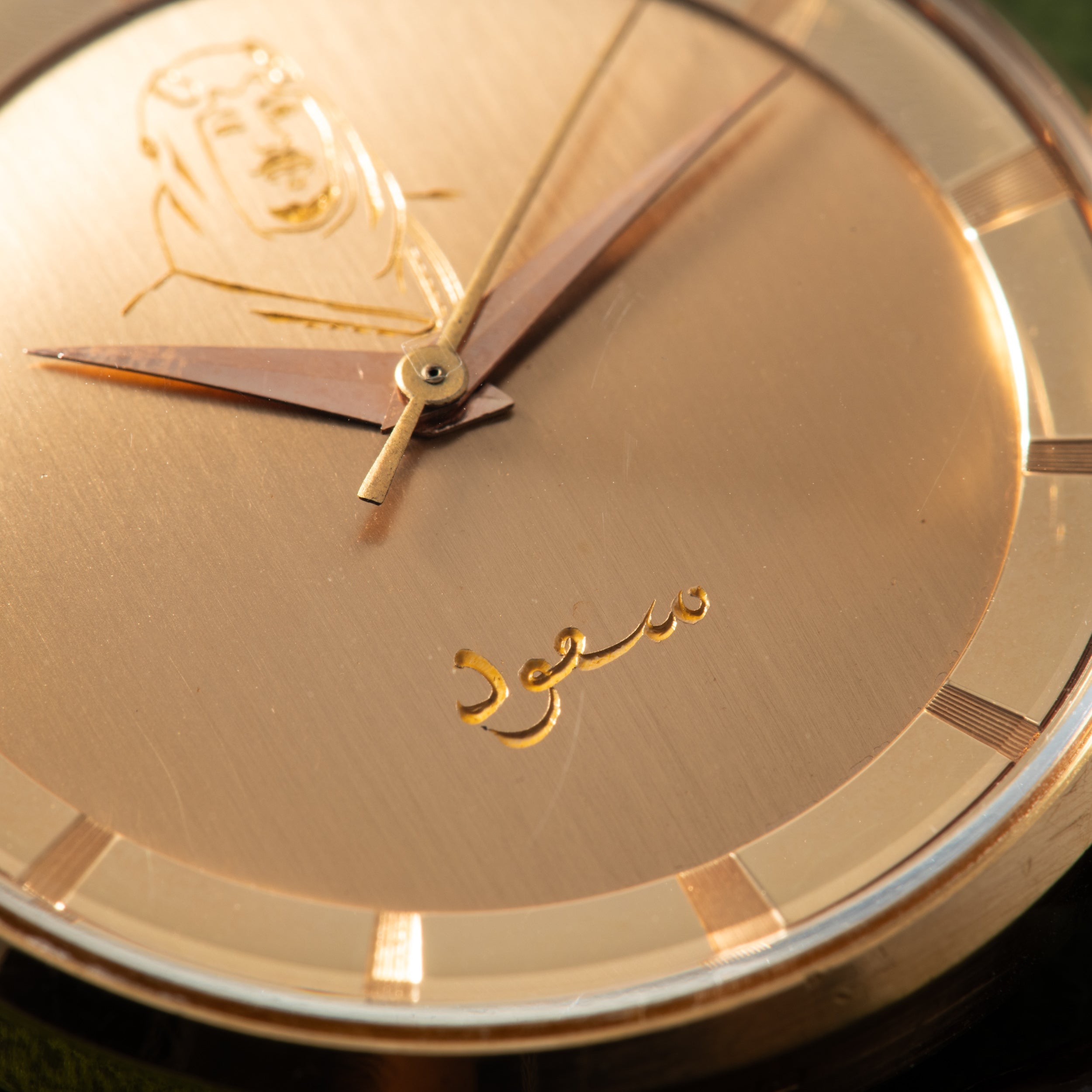 Gold watch dial with portrait engraving, Arabic script, and gold hands on brushed surface.