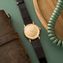Gold-toned round watch with dark leather strap, displayed with brown leather pouch and green beaded bracelet on teal backgrou