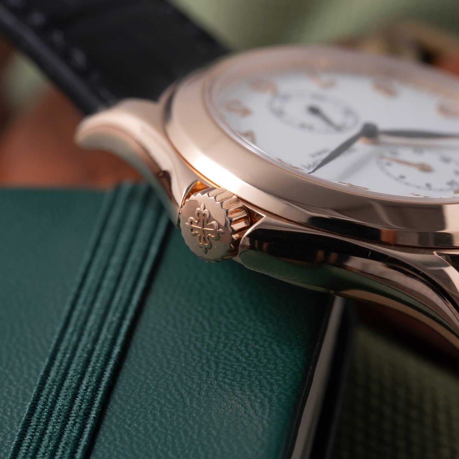 Rose gold watch with white dial, Arabic numerals, and crown with emblem detail, resting on green leather.