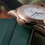 Rose gold watch with white dial, Arabic numerals, and crown with emblem detail, resting on green leather.