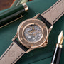 Rose gold watch with skeletonized dial showing mechanical movement, black leather strap, displayed on green fabric with pen.
