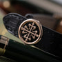 Rose gold watch with ornate cross motif dial, black leather strap, displayed at an angle.