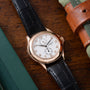 Rose gold watch with cream dial, Arabic numerals, black leather strap, and two subdials on wooden surface with leather access