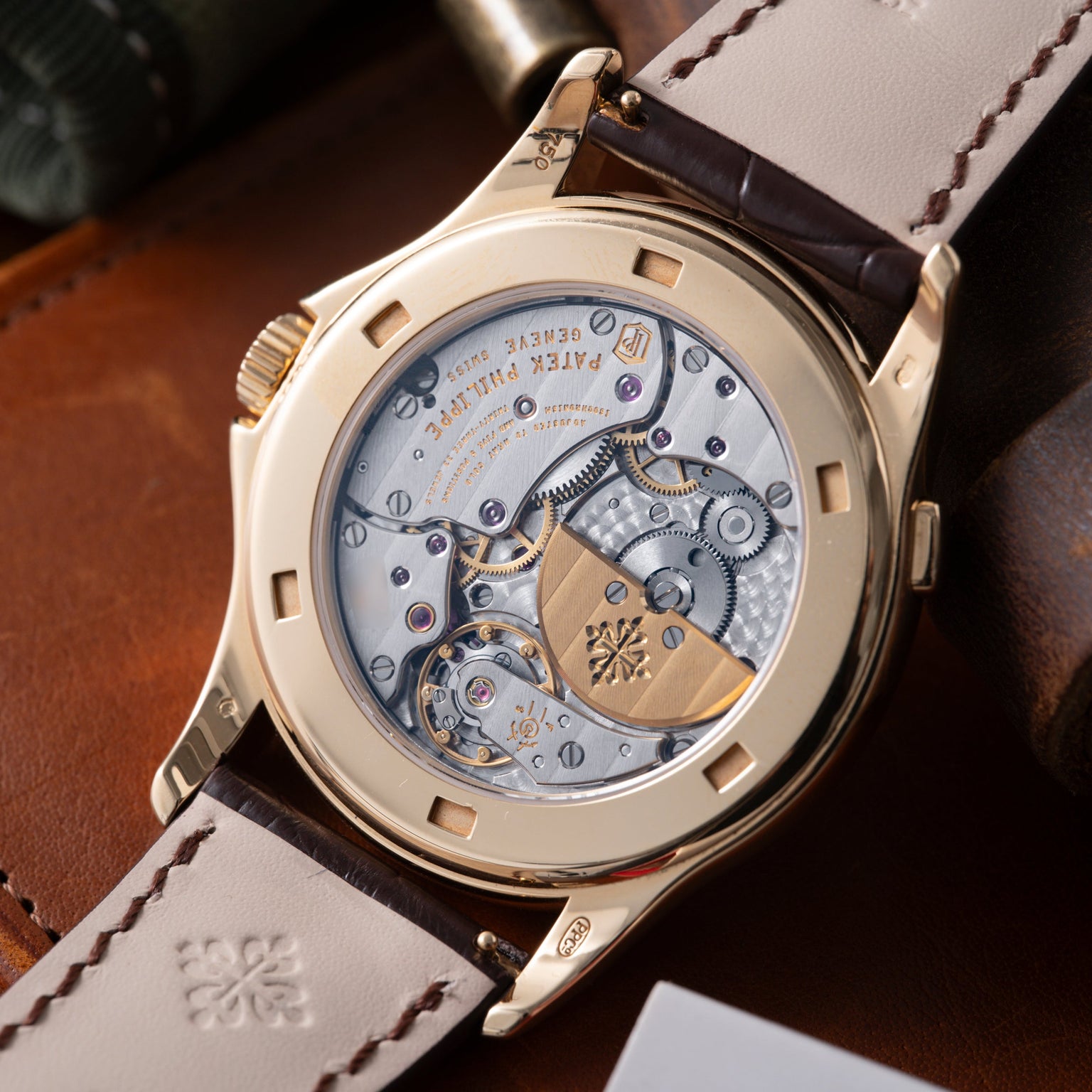 Gold-tone watch with exposed movement visible through caseback, dark leather strap with contrast stitching.