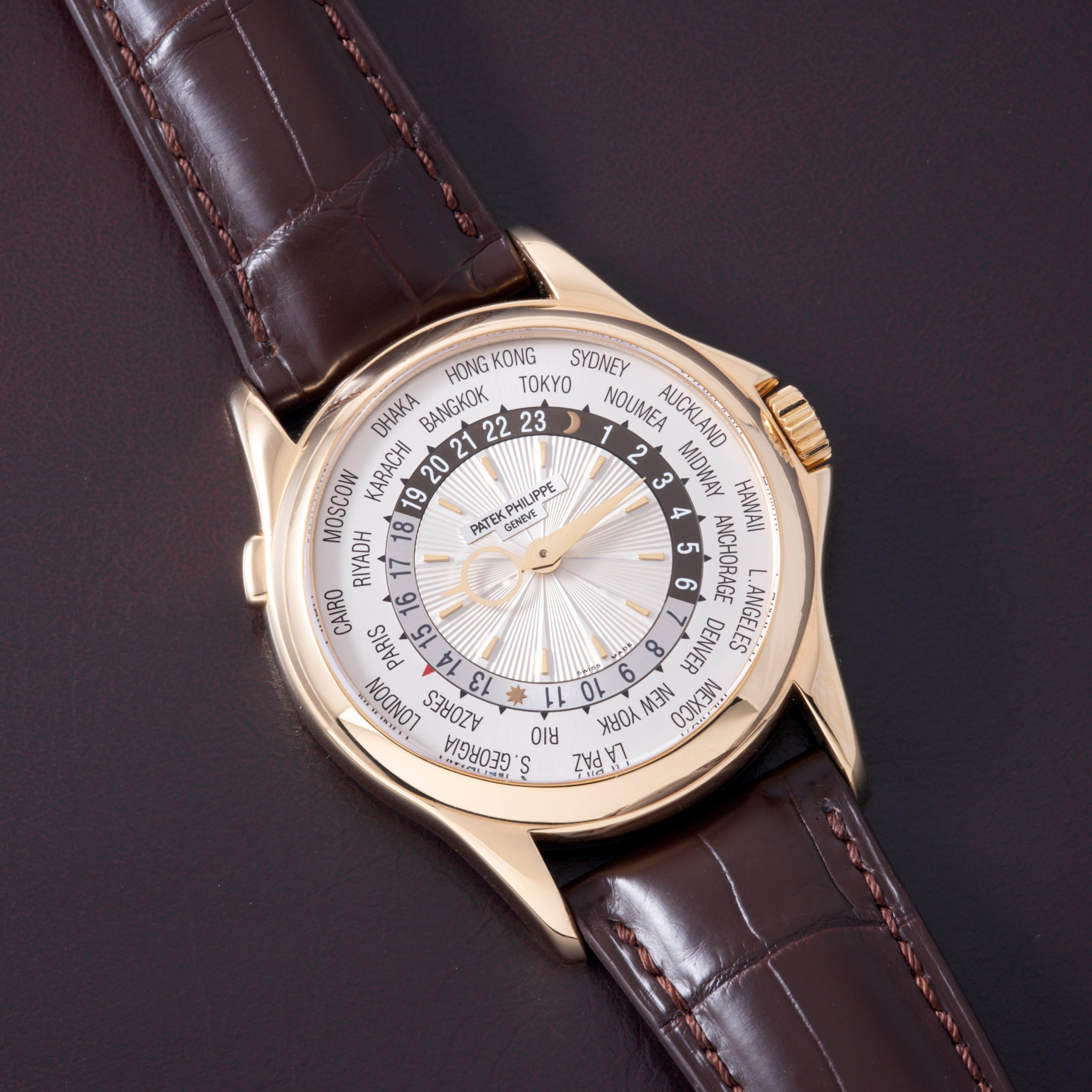 Gold watch with white dial, world city names around outer ring, dark leather crocodile-textured strap with orange stitching.