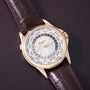 Gold watch with white dial, world city names around outer ring, dark leather crocodile-textured strap with orange stitching.
