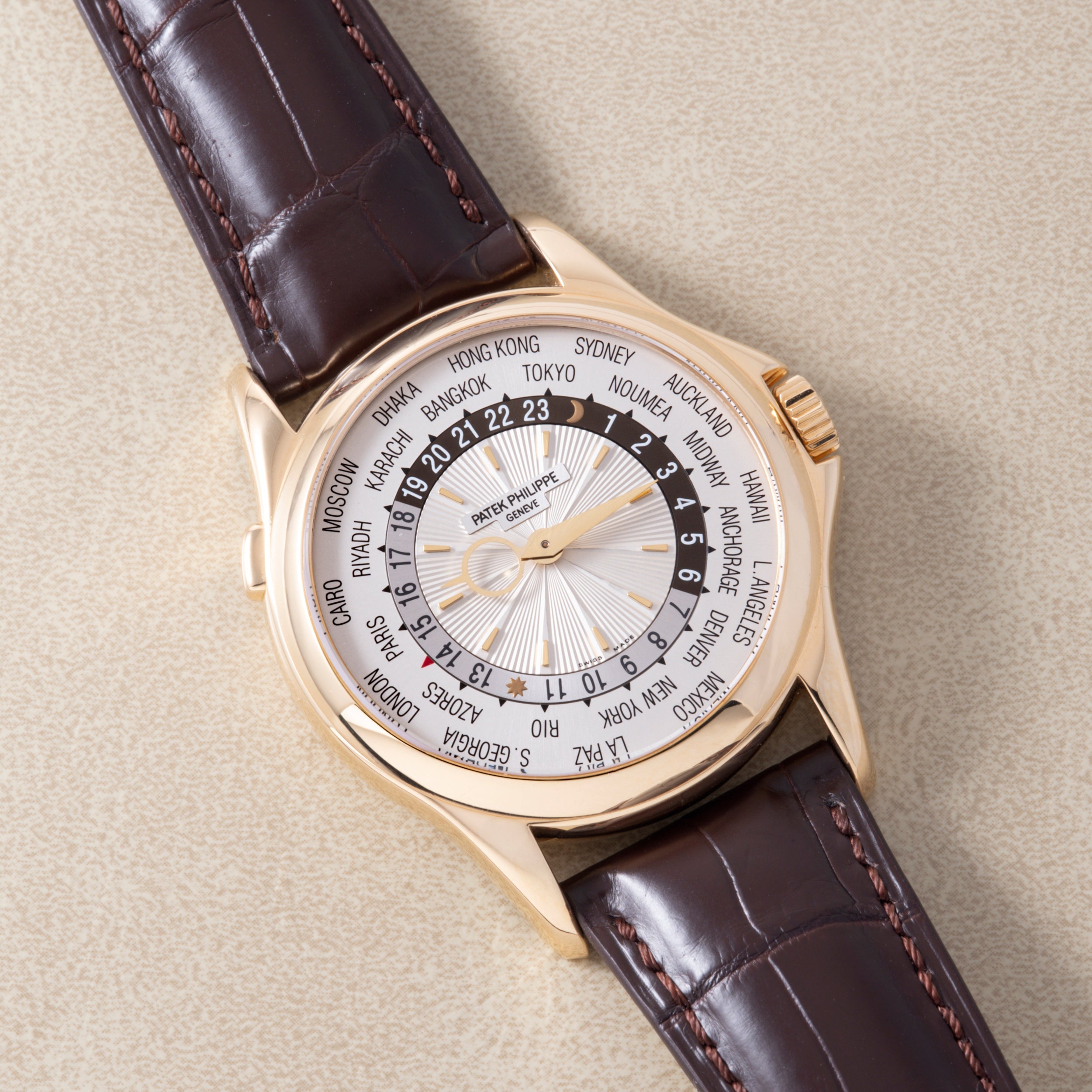 Gold-tone watch with world time zone dial and brown leather strap on neutral background.