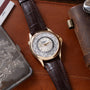 Gold-tone watch with white dial displaying world city names and 24-hour time zone ring, dark brown leather strap.