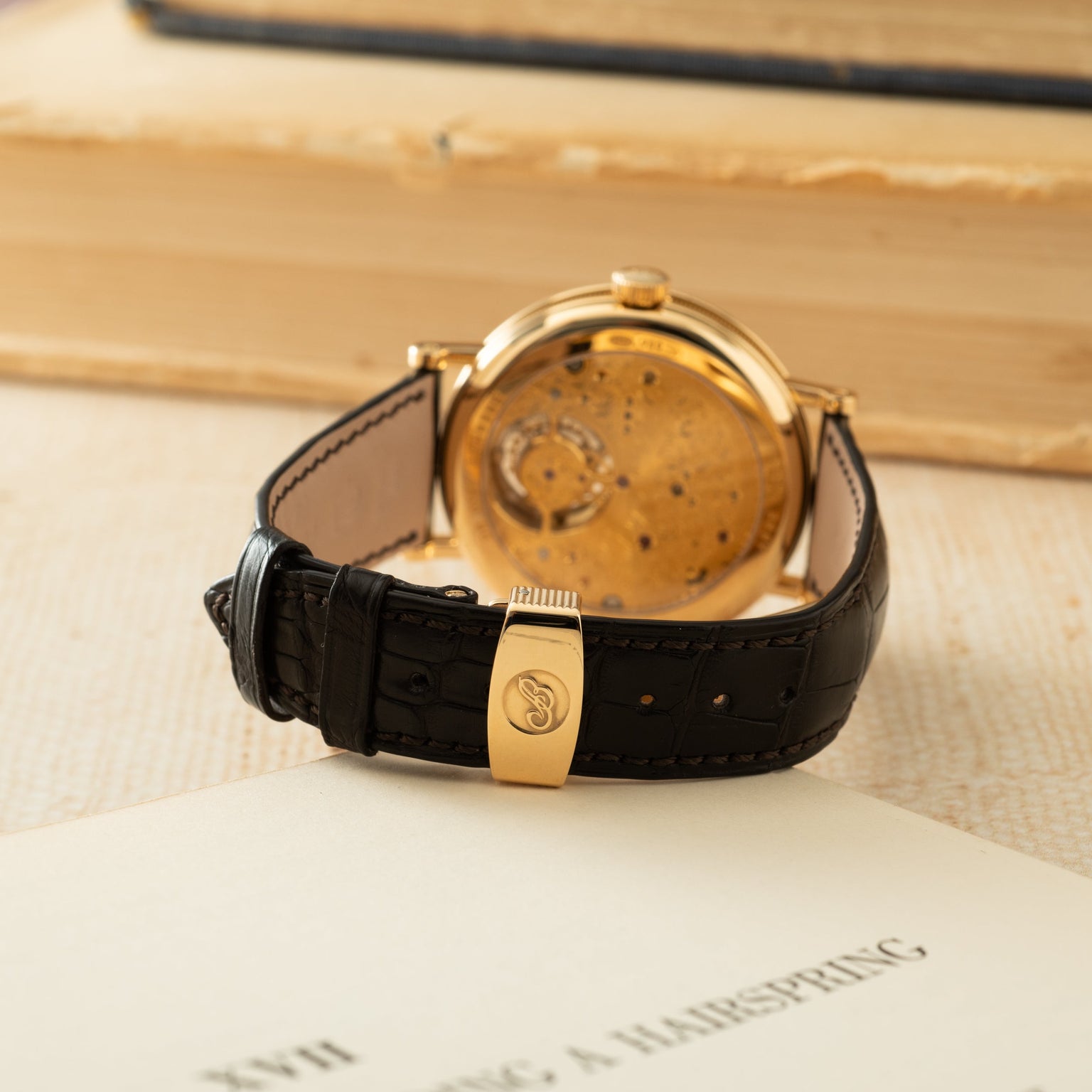 Gold-toned watch with visible tourbillon mechanism, black crocodile leather strap, and gold clasp displayed caseback-up.