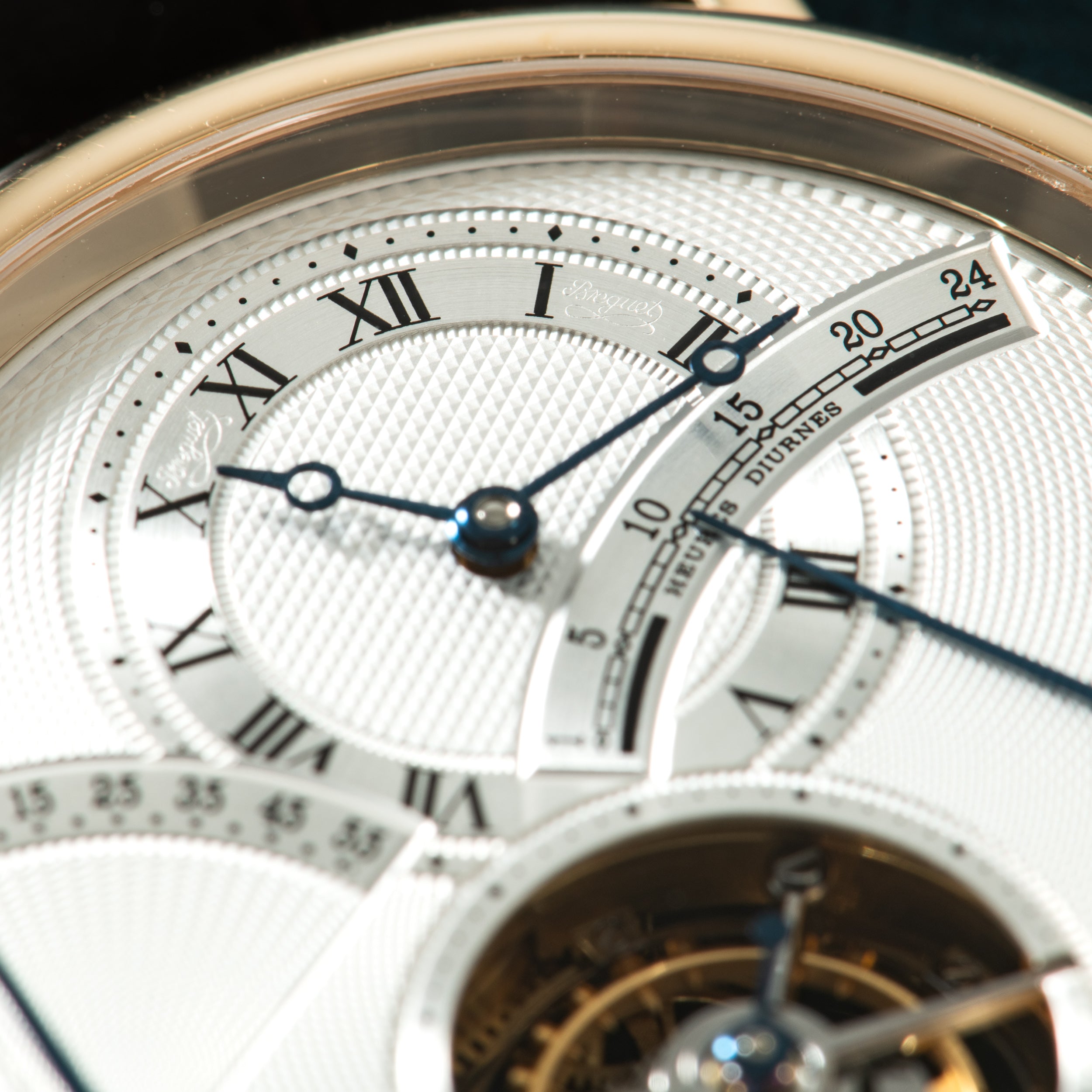 Gold-tone watch face with white dial, Roman numerals, blued hands, date window, and tourbillon visible at bottom.