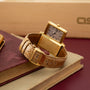 Gold rectangular watch with cream dial and Arabic numerals on tan crocodile leather strap, displayed on burgundy fabric.
