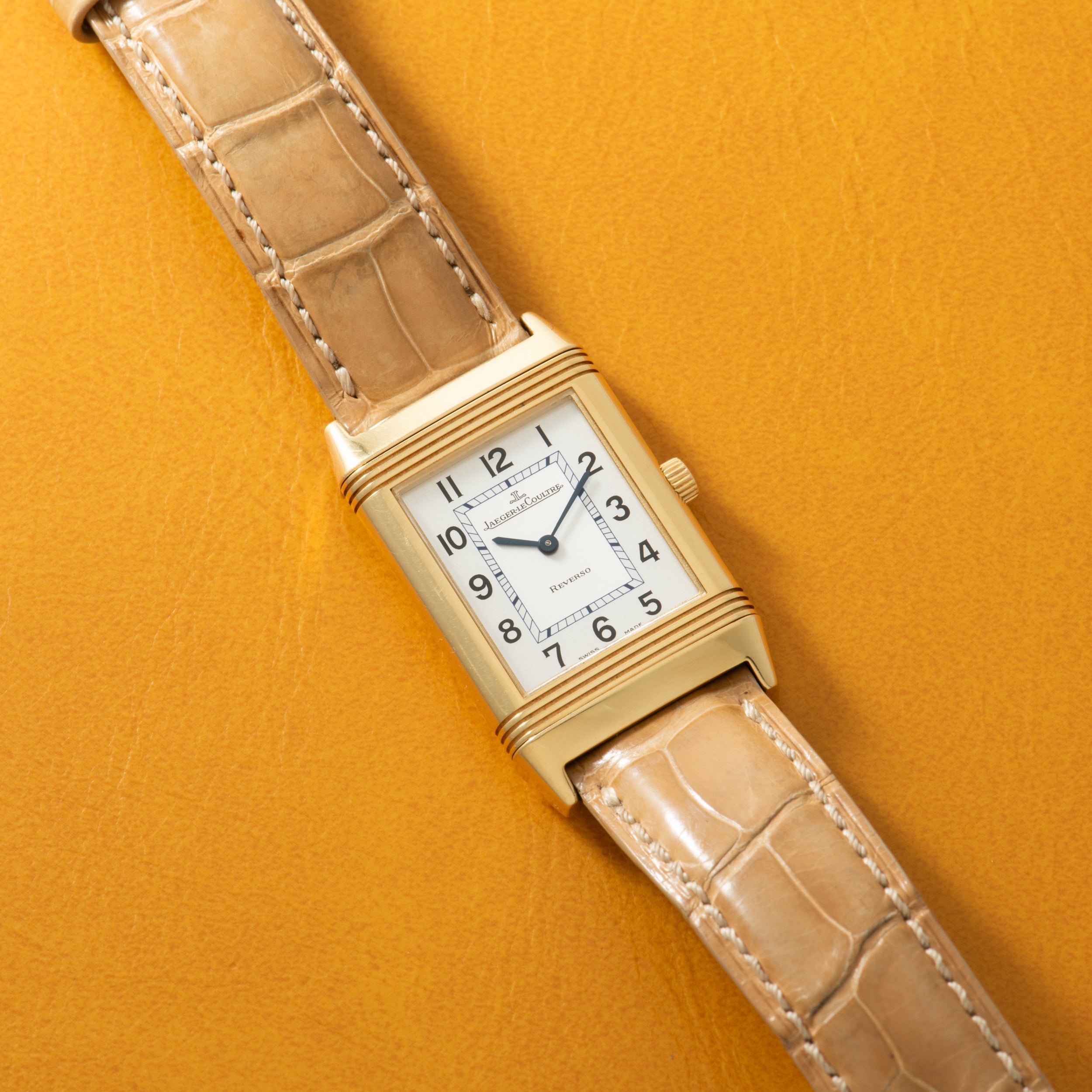 Gold-tone rectangular watch with white dial, Arabic numerals, and tan crocodile leather strap on yellow background.