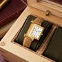 Gold-tone rectangular watch with white dial and brown leather strap, displayed in wooden box with olive fabric interior.