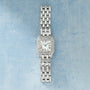 Stainless steel rectangular watch with diamond-set bezel, cream dial with Roman numerals, blue hands, and multi-link metal br