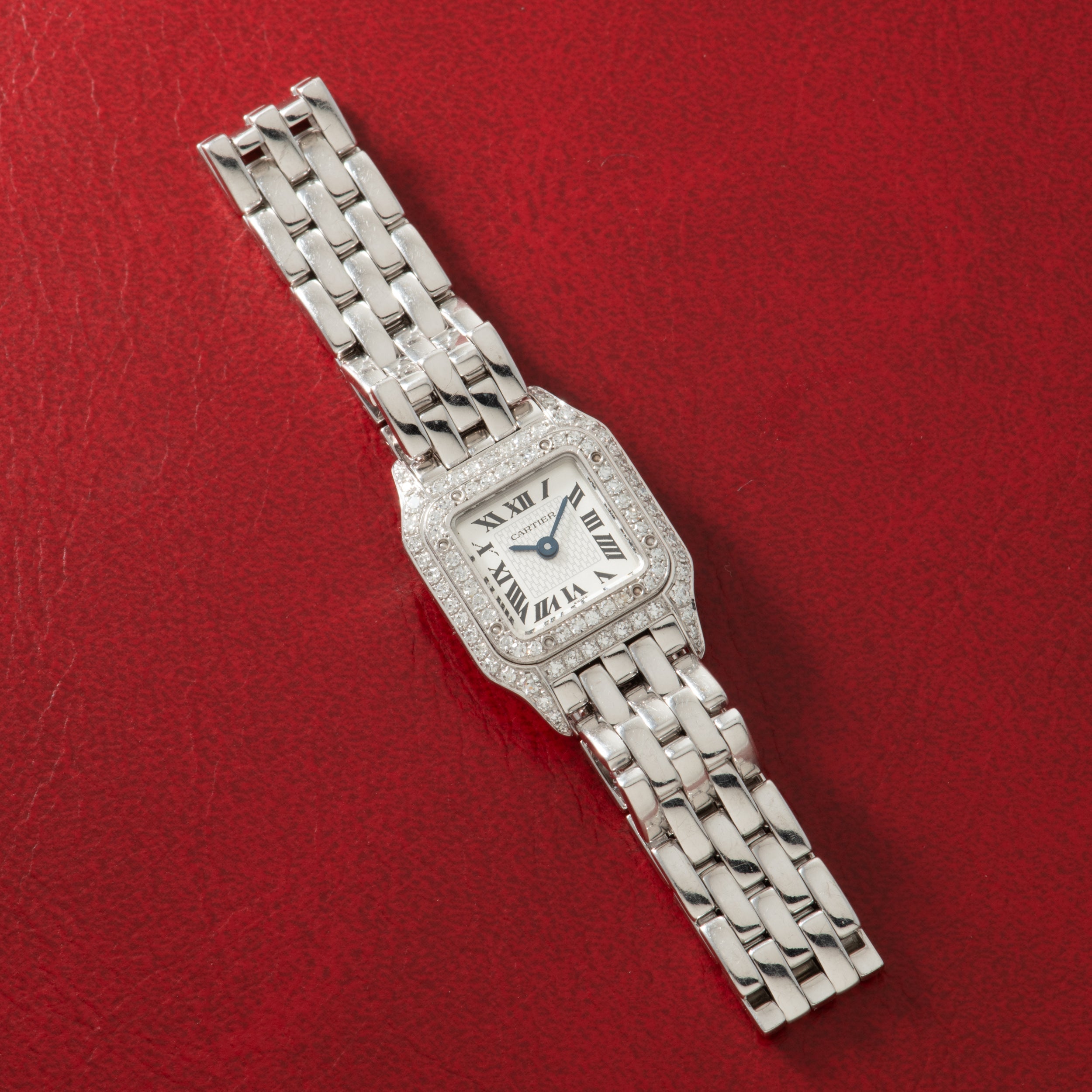 Silver-toned watch with square face, diamond-set bezel, Roman numerals, and matching metal bracelet on red background.