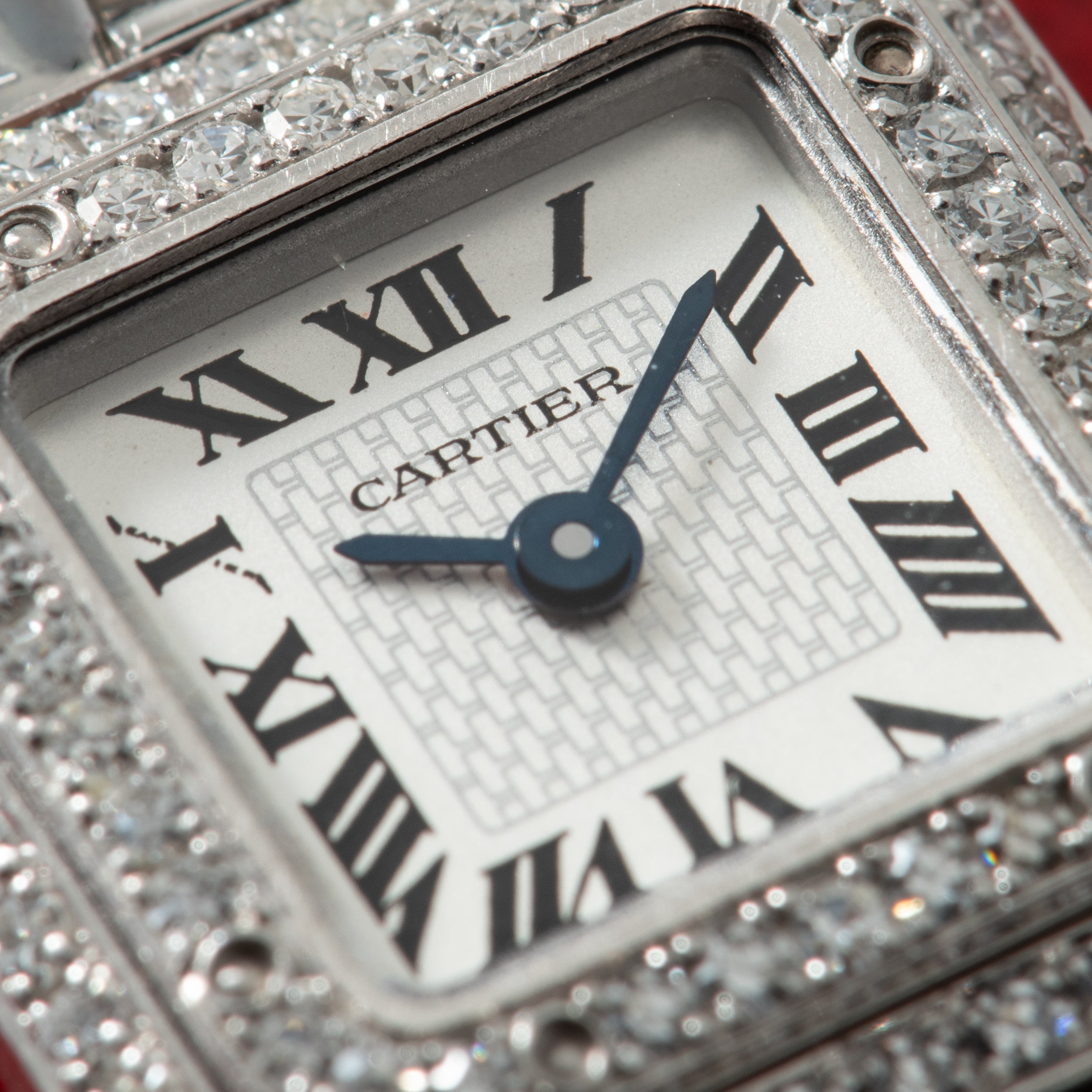 Cartier watch face with Roman numerals and blue hands, surrounded by diamond-set bezel on metal case.