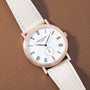 Rose gold watch with white dial, Roman numerals, subdial, and white leather strap on tan background.