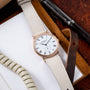 Rose gold watch with white dial, Roman numerals, and cream leather strap displayed on brown leather accessory.