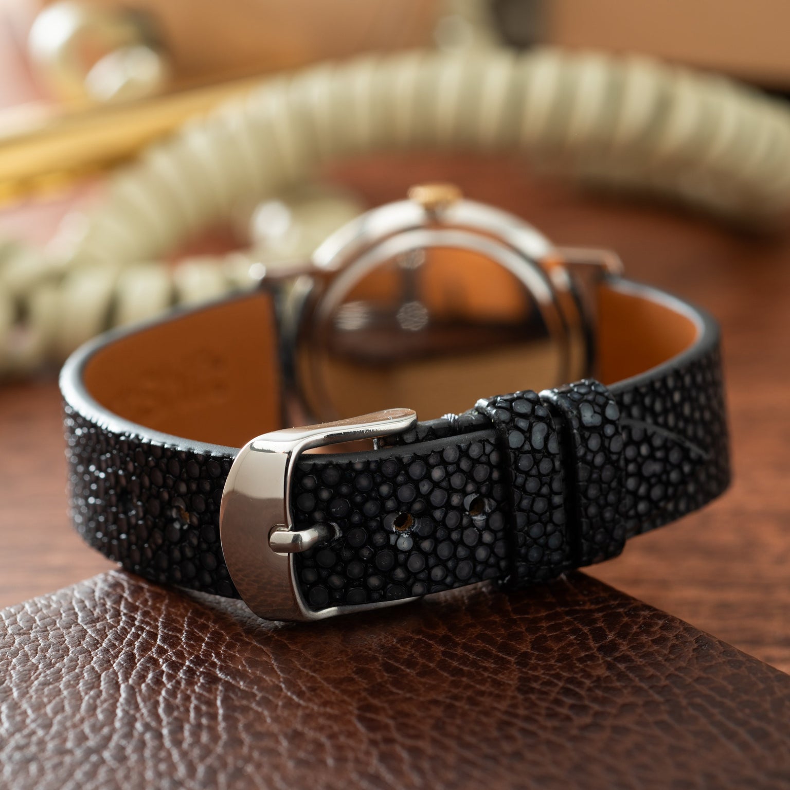 Black textured leather strap watch with silver-tone case and tan dial displayed on brown surface.