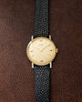 Hermès watch with gold-tone round case, cream dial, and black textured leather strap on brown leather background.