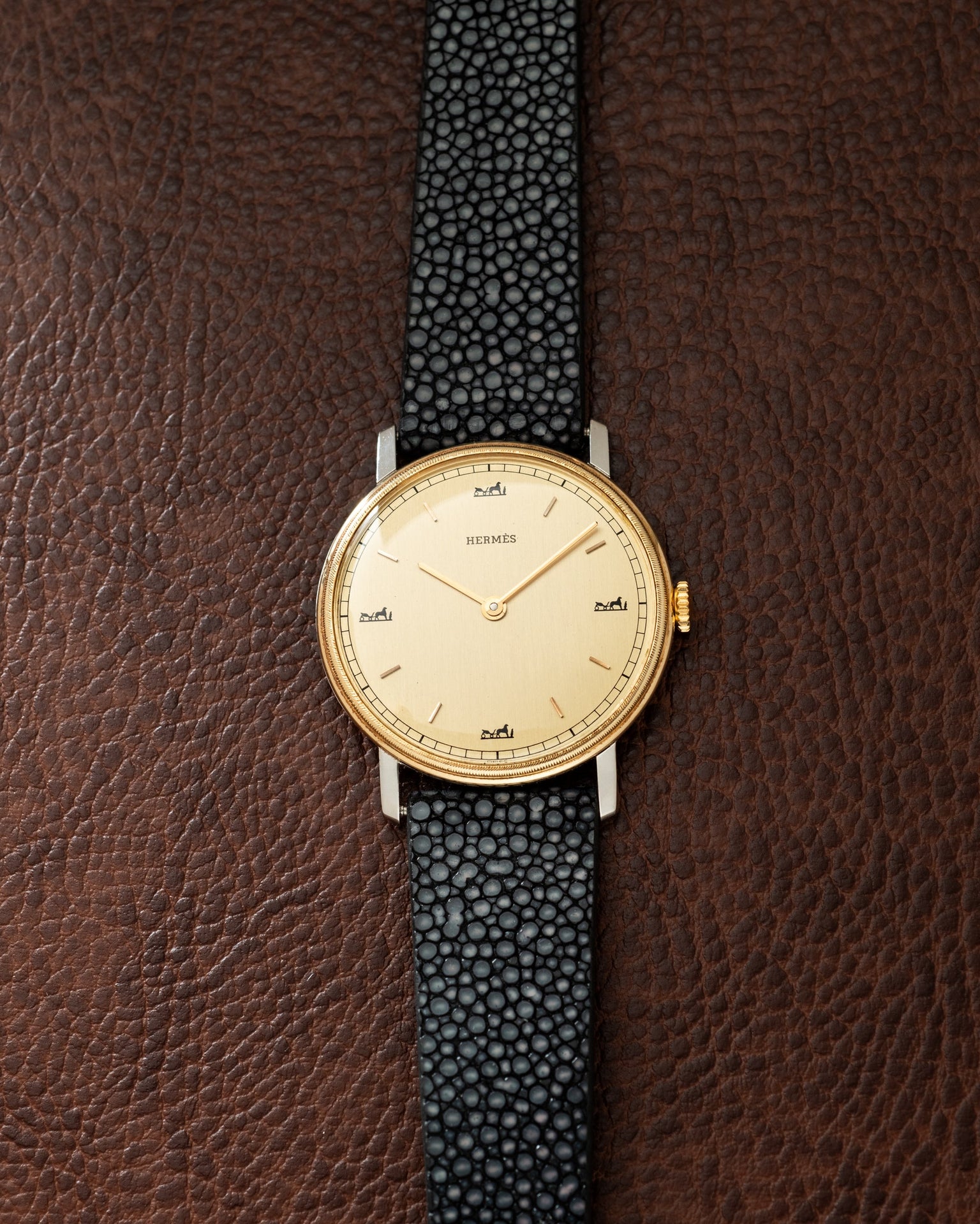 Hermès watch with gold-tone round case, cream dial, and black textured leather strap on brown leather background.