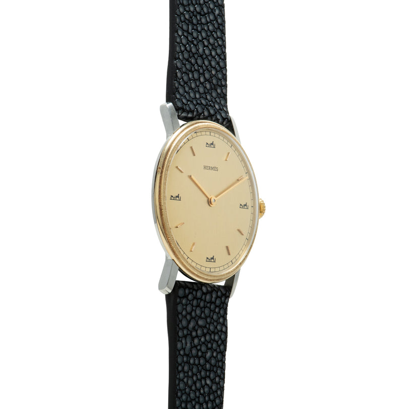 Hermès wristwatch with oval cream dial, gold-tone hands and indices, two-tone case, and black textured leather strap.
