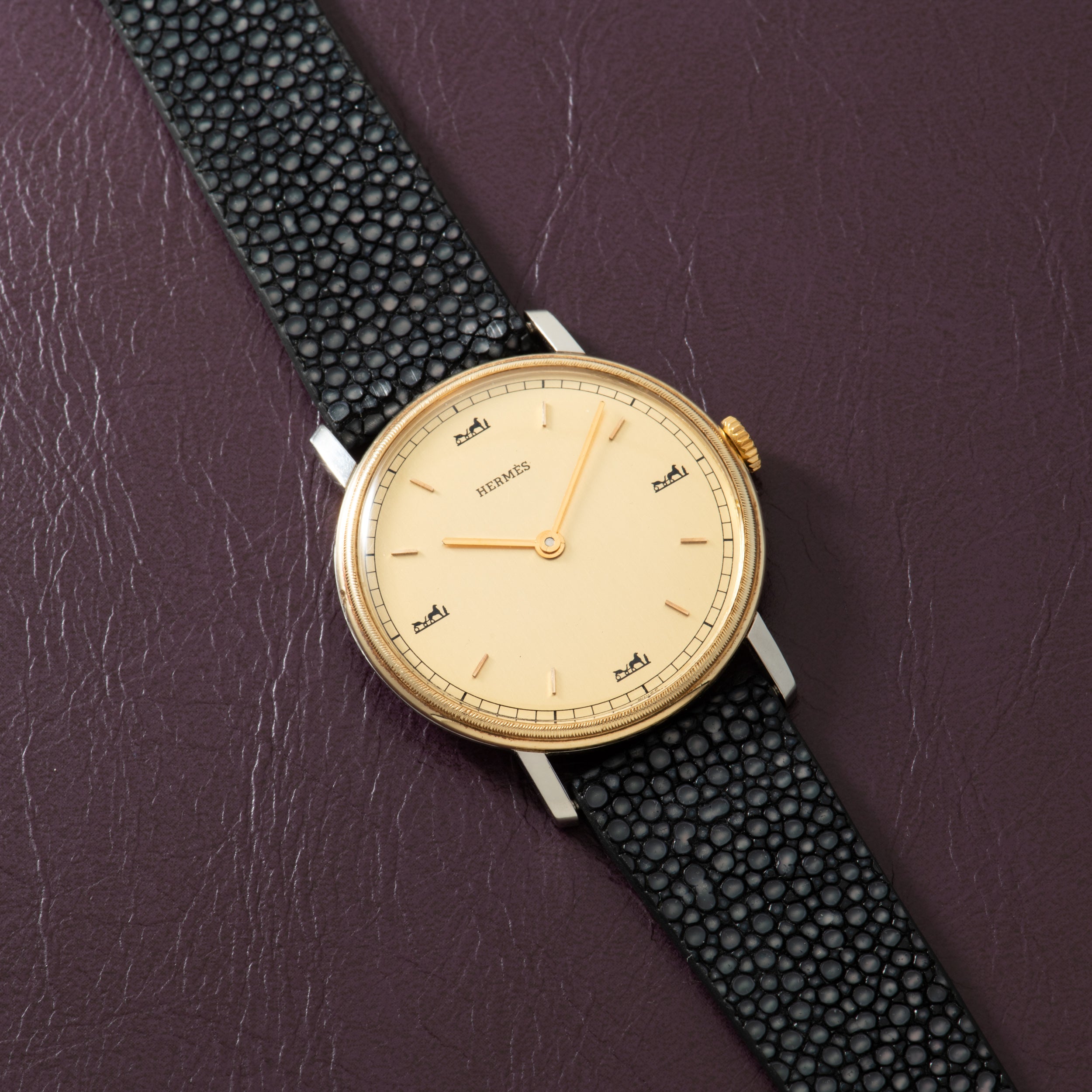 Hermès watch with cream dial, gold-tone case, Arabic numerals, and black textured leather strap on purple background.