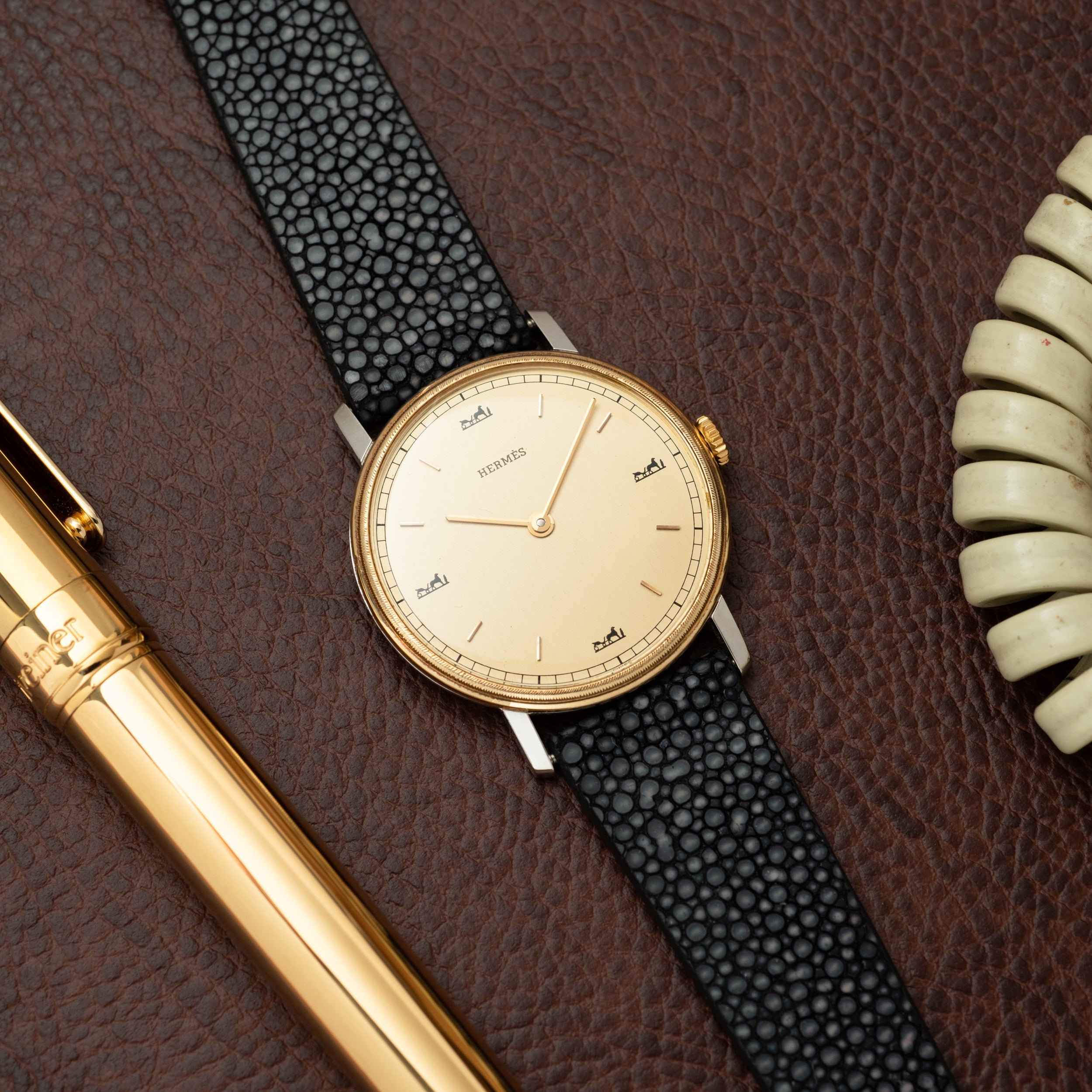 Hermès watch with cream dial, gold-tone case, and black textured leather strap on brown leather background.