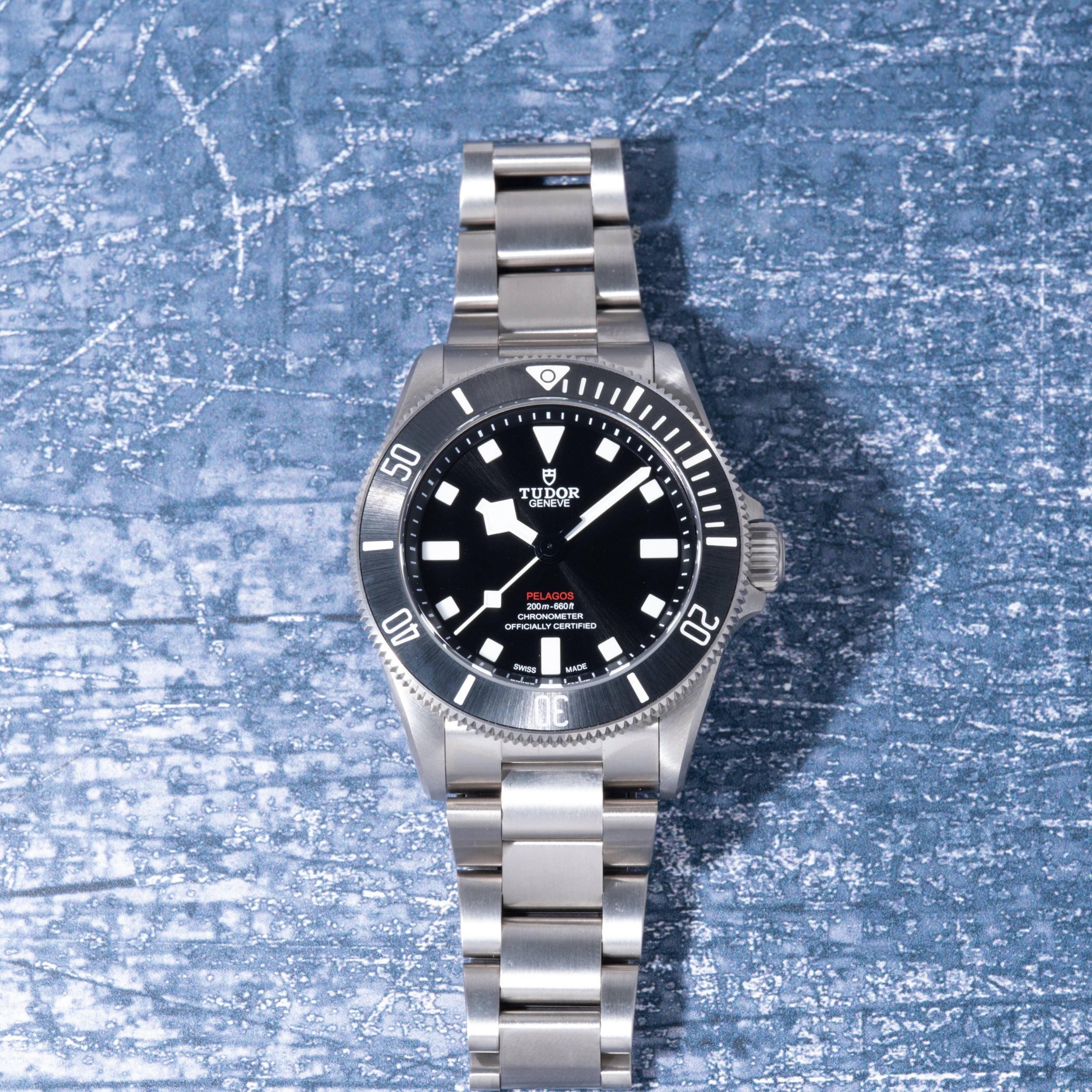 Tudor Pelagos 39 stainless steel dive watch with black dial, white hour markers, and matching metal bracelet on textured blue