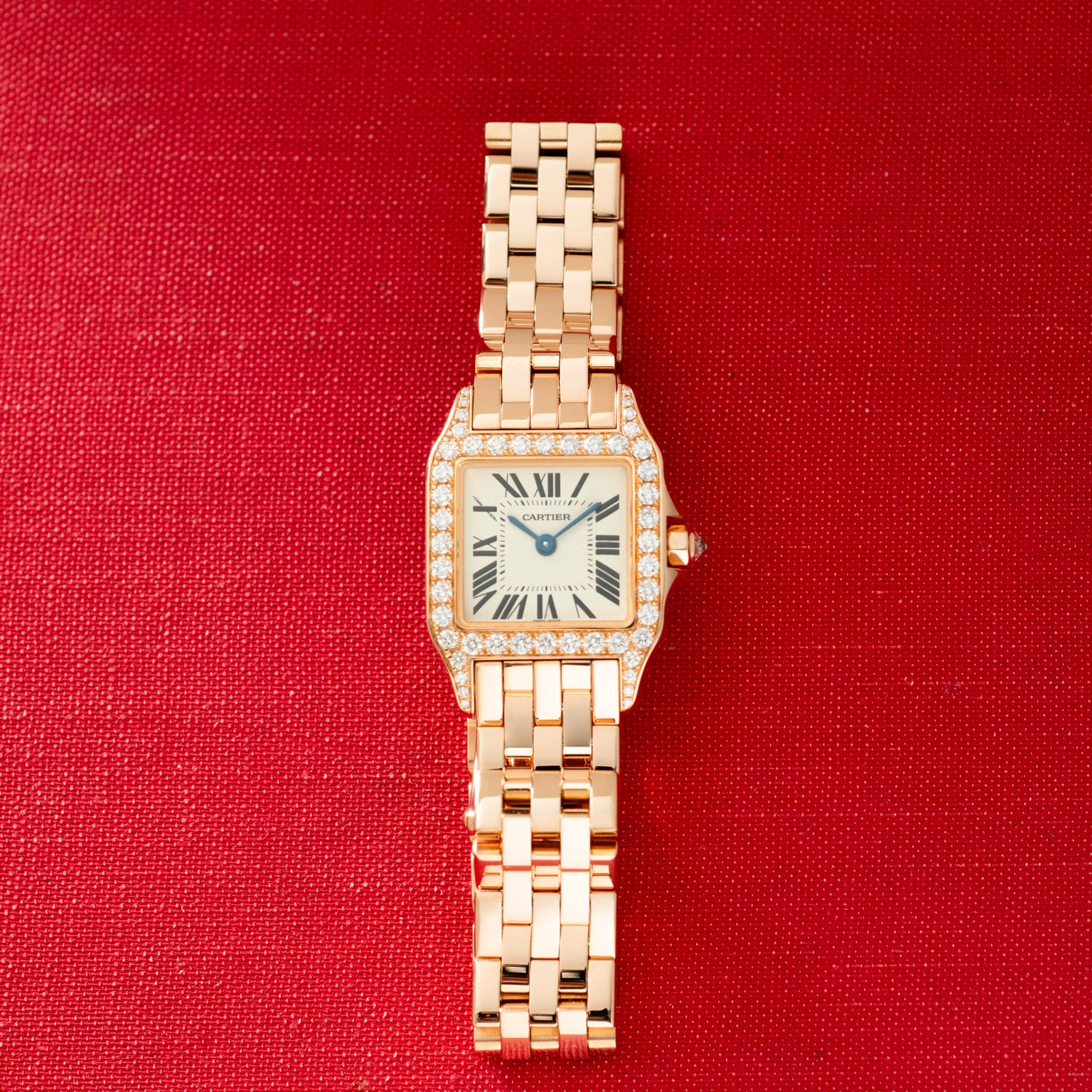 Rose gold watch with square face, diamond-set bezel, Roman numeral dial, and linked bracelet on red background.
