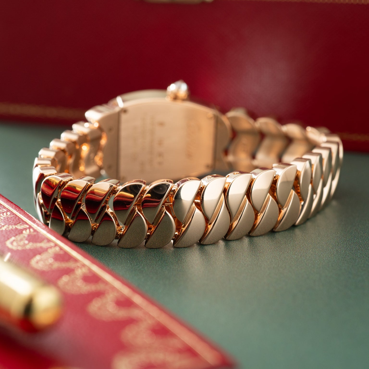 Gold and stainless steel bracelet watch with segmented links and rectangular face on red and green background.