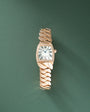 Rose gold watch with rectangular white dial, Roman numerals, diamond-set bezel, and intertwined rose gold bracelet against gr