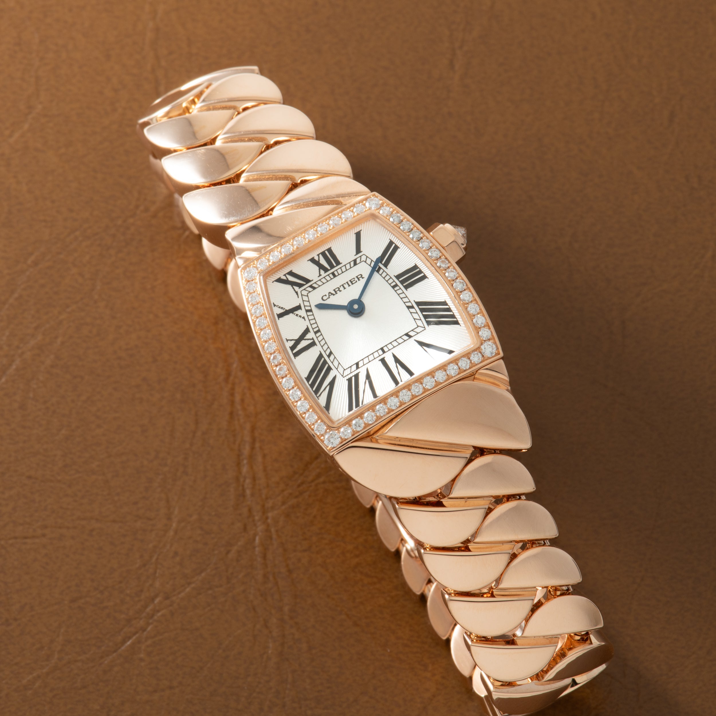 Rose gold watch with tonneau-shaped white dial, Roman numerals, diamond-set bezel, and articulated scalloped bracelet.