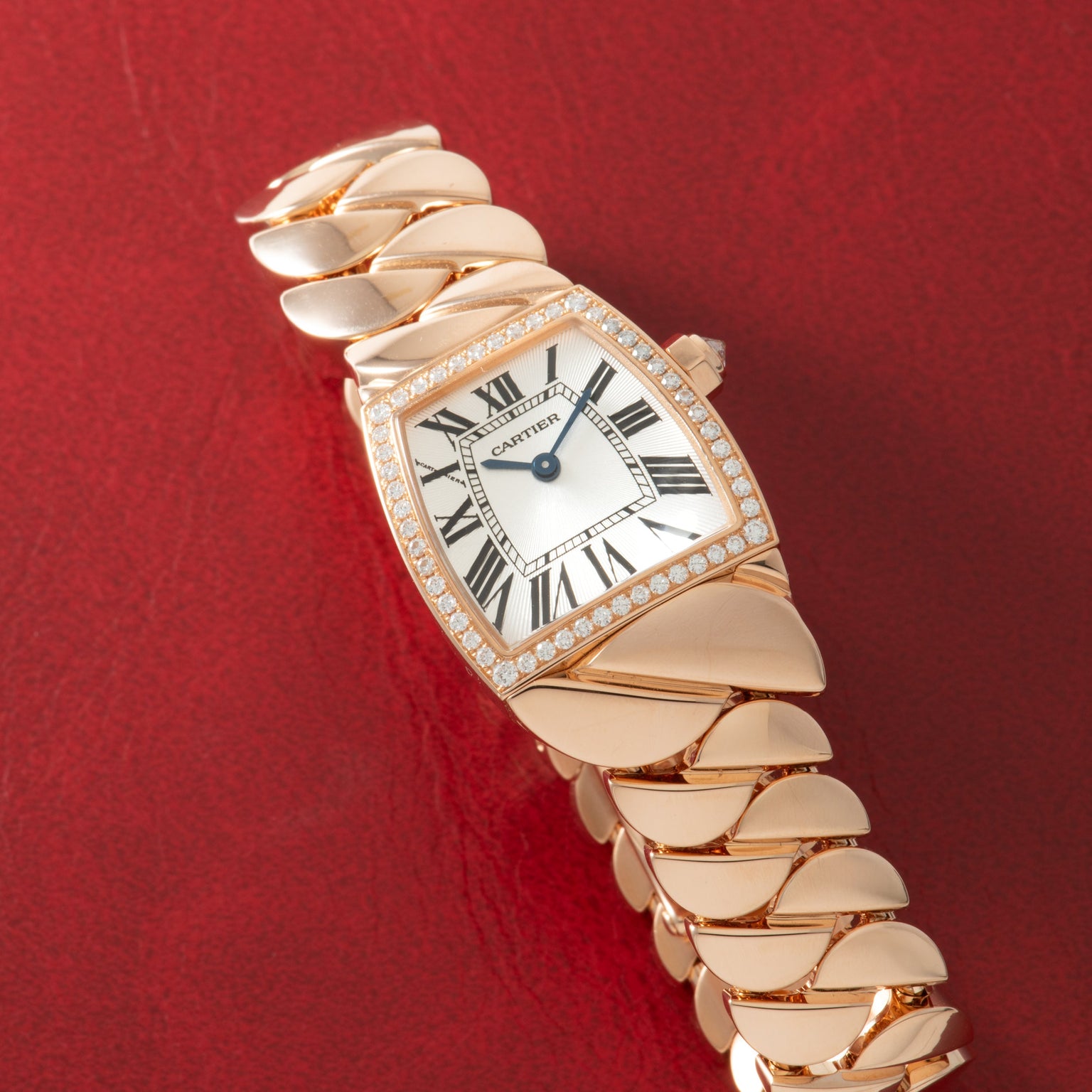 Rose gold Cartier watch with tonneau-shaped dial, diamond bezel, Roman numerals, and segmented bracelet on red background.