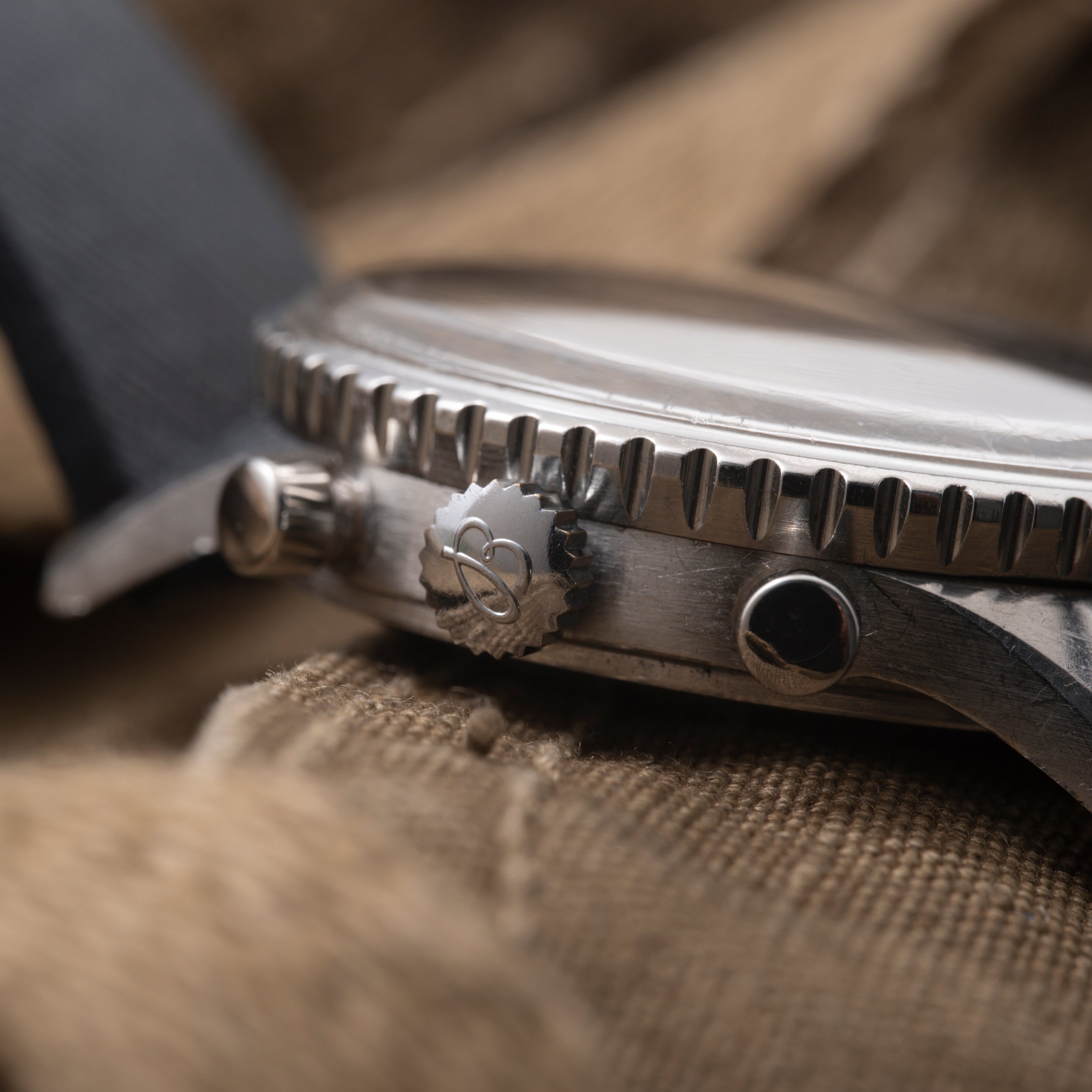 Steel watch with fluted bezel, crown with logo, and dark dial visible from side angle on tan fabric.