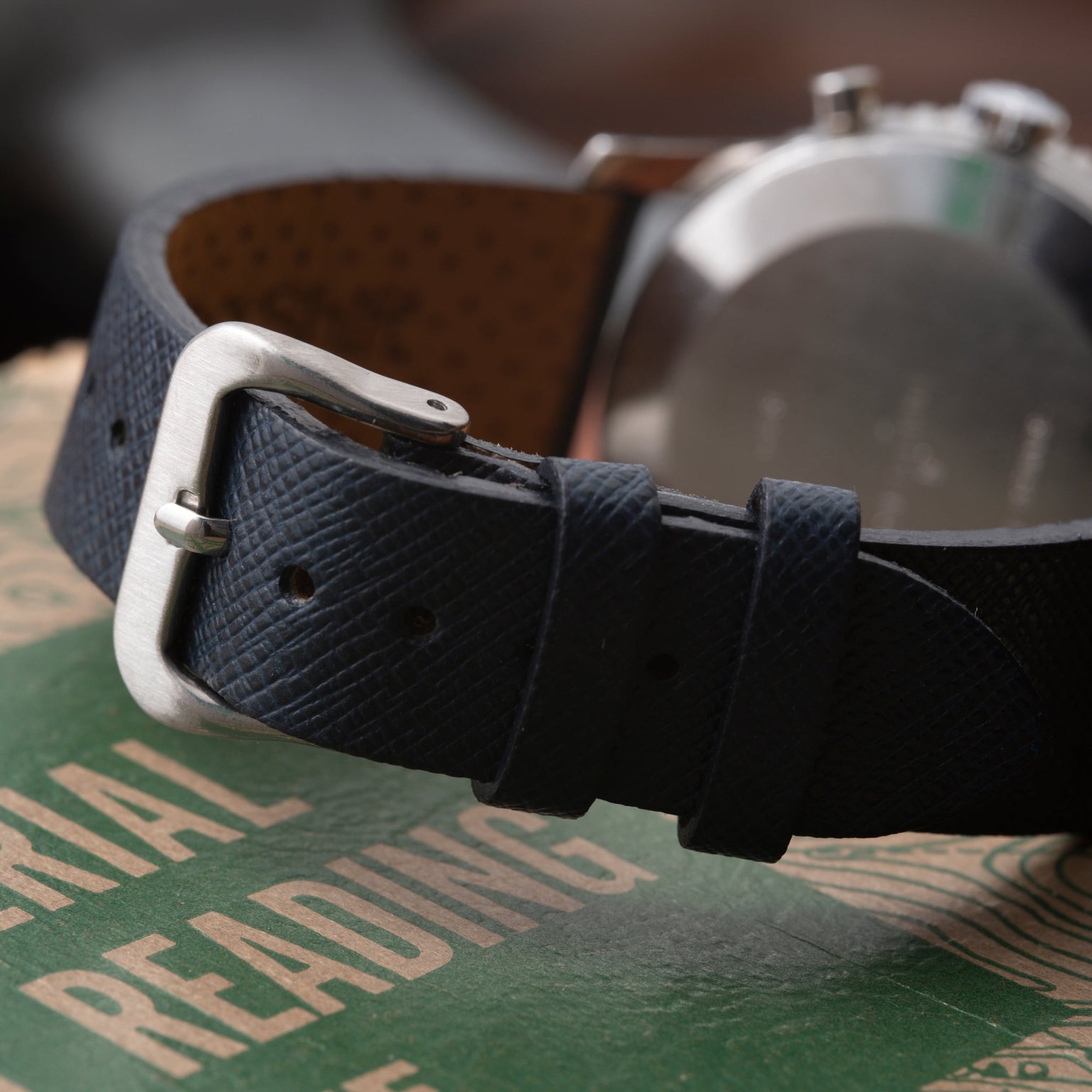 Stainless steel watch with dark blue leather strap and silver buckle, displayed on green branded packaging.