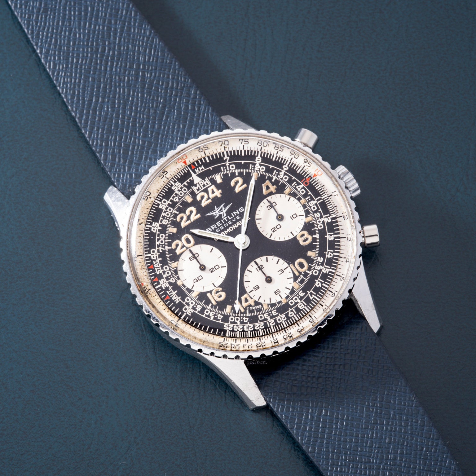 Silver chronograph watch with black dial, 24-hour subdials, and gray fabric strap on blue background.