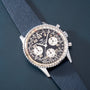 Silver chronograph watch with black dial, 24-hour subdials, and gray fabric strap on blue background.
