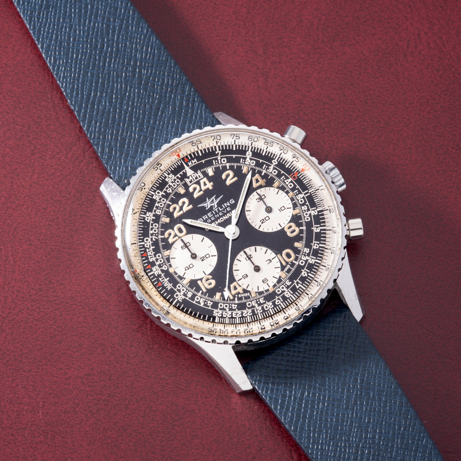 Breitling Cosmonaute chronograph watch with black dial, white subdials, and blue fabric strap on burgundy background.